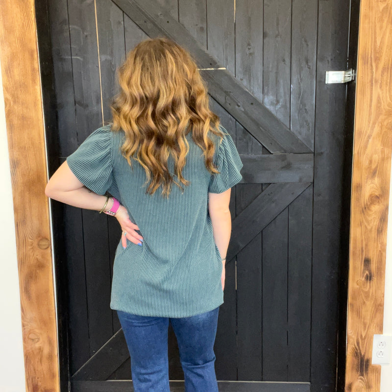 Person wearing a teal top and blue jeans standing in front of a wooden door.