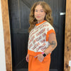 Woman wearing a 'GAMEDAY' sweater and orange pants standing in front of a wooden door.