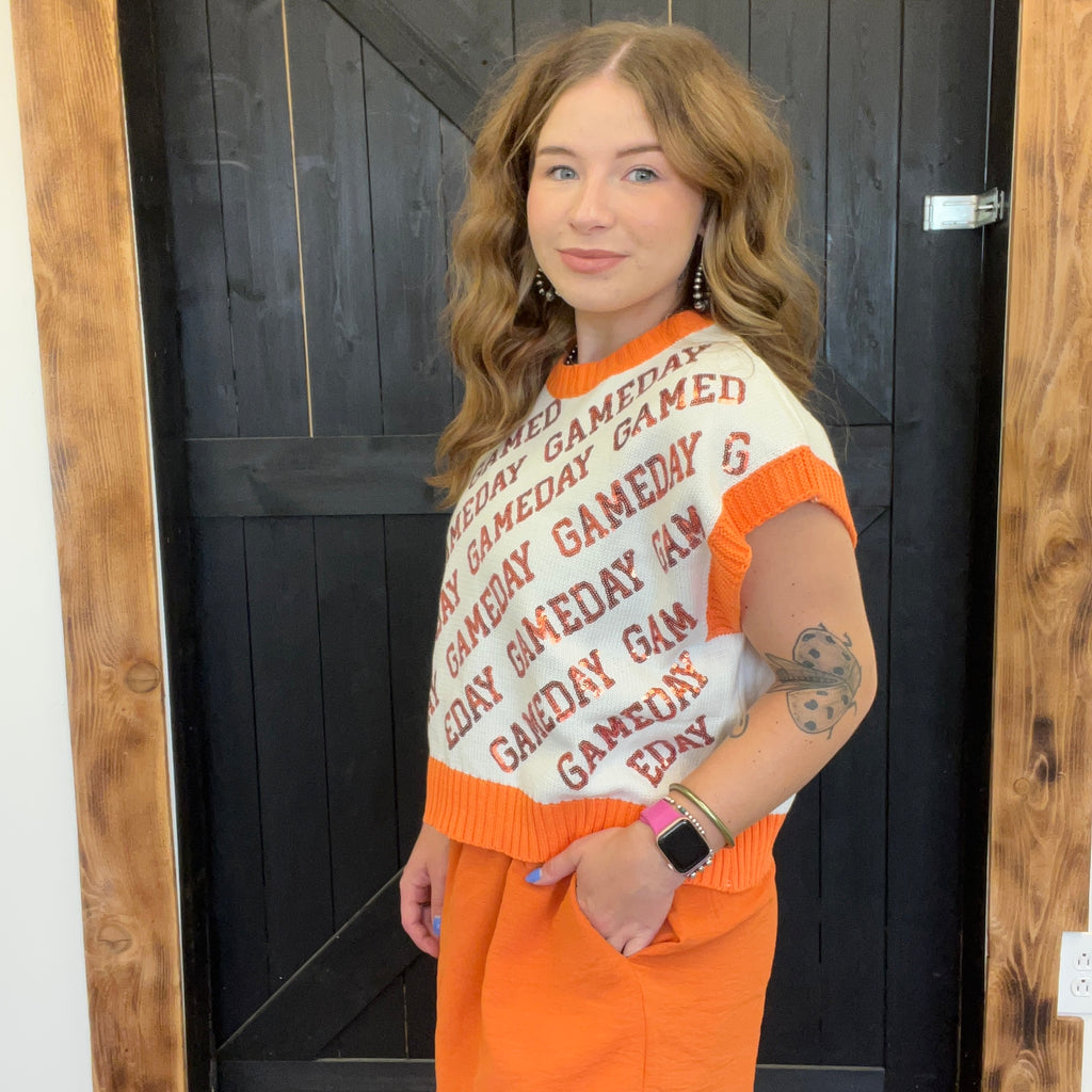 Woman wearing a 'GAMEDAY' sweater and orange pants standing in front of a wooden door.