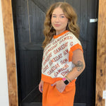 Woman wearing a 'GAMEDAY' sweater and orange pants standing in front of a wooden door.