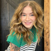 Woman with wavy hair wearing a green sweater over a striped shirt, standing against a wooden and dark wall.