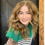 Woman with wavy hair wearing a green sweater over a striped shirt, standing against a wooden and dark wall.