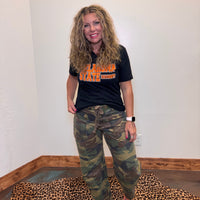 Person wearing a black t-shirt with orange text and camouflage pants standing in a room.