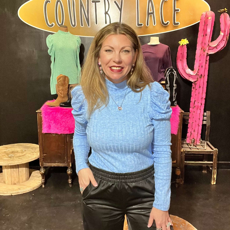 A person wearing a blue long sleeve top with a mock-neck neckline and puffed sleeves, standing in front of a display with pink mannequins.