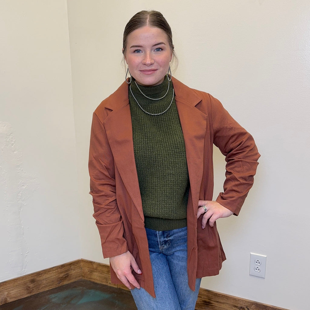 Woman wearing a brown jacket over a black top and light blue jeans, standing against a plain wall.