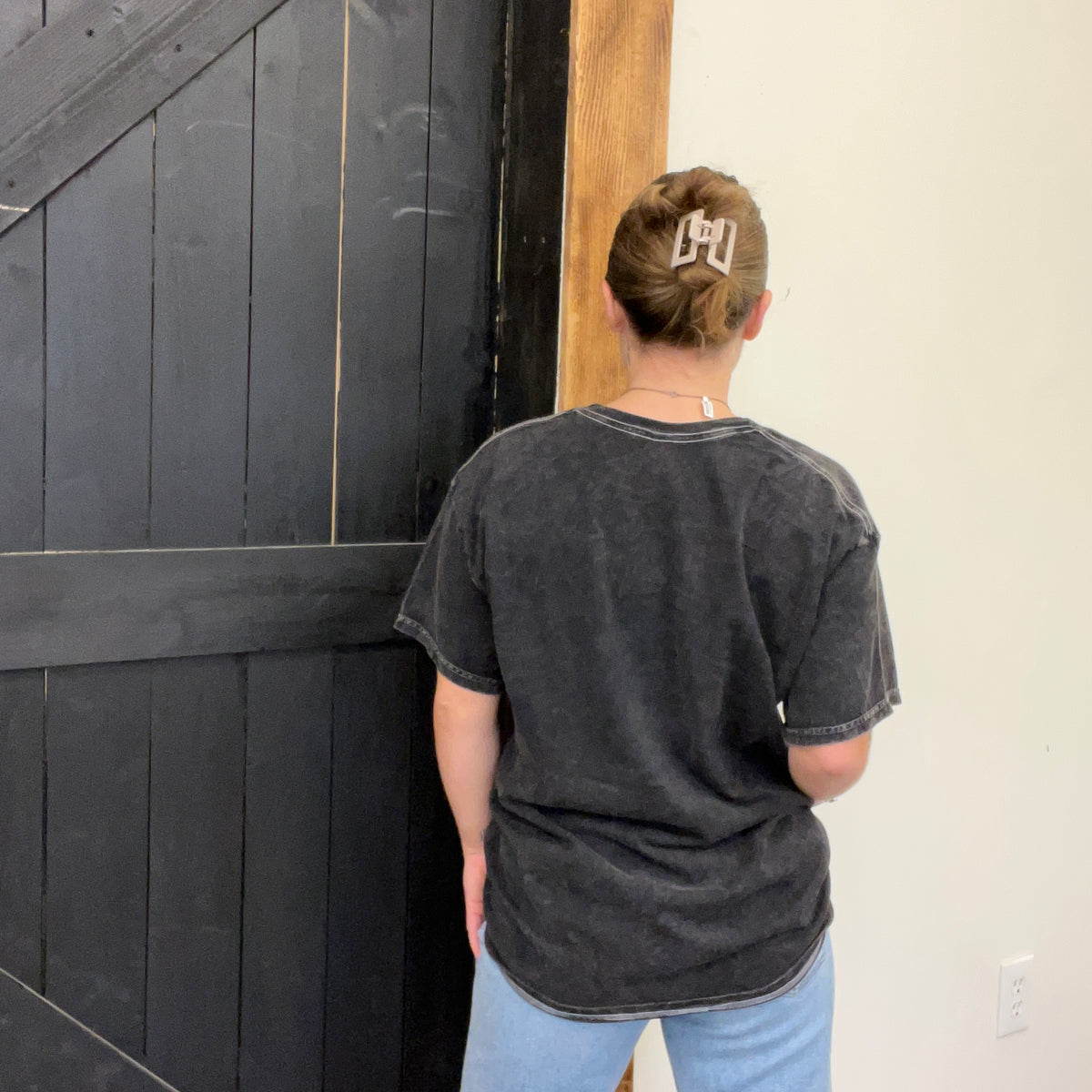 Person wearing a dark gray t-shirt with a logo, standing in front of a black wooden door.