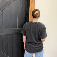 Person wearing a dark gray t-shirt with a logo, standing in front of a black wooden door.
