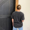 Person wearing a dark gray t-shirt with a logo, standing in front of a black wooden door.