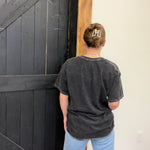 Person wearing a dark gray t-shirt with a logo, standing in front of a black wooden door.