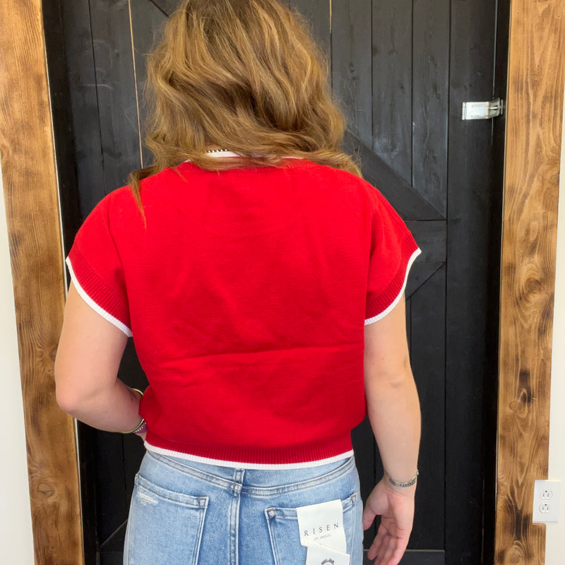 Person wearing a red sweater and blue jeans with a tag, standing in front of a wooden door.