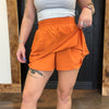 Person wearing orange shorts and a white top, standing indoors with wooden elements.