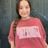 Girl wearing a red 'University of Oklahoma' shirt standing against a wooden background