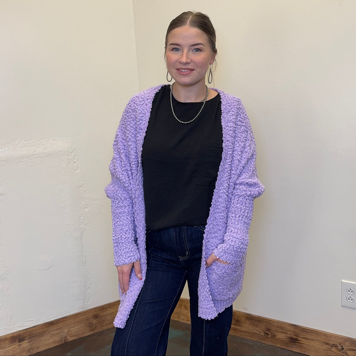 Woman wearing a purple cardigan over a black top in a room with a white wall and wooden trim.