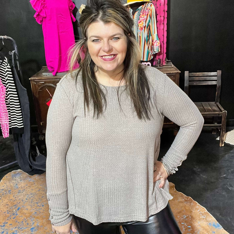 A woman smiling, wearing a long sleeve top in a beige color, with a wide neckline, in an indoor setting with clothes in the background.
