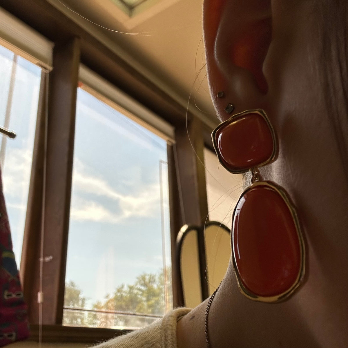Close-up of a person wearing large red earrings with gold frames, sitting by a window.
