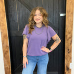Woman wearing a purple top and blue jeans standing in front of a wooden door.