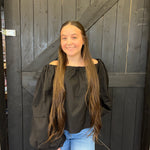 Young woman with long hair wearing a black top and blue jeans standing in front of a wooden door.
