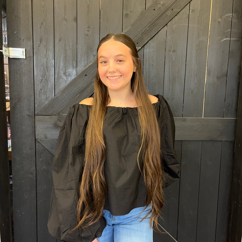 Young woman with long hair wearing a black top and blue jeans standing in front of a wooden door.