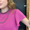 Person wearing a pink top and black and white necklace, standing in front of a wooden door.