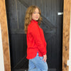Woman in a red jacket and blue jeans standing in front of a black wooden door.