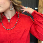 Person wearing a red shirt with a necklace and jewelry, standing against a wooden door.