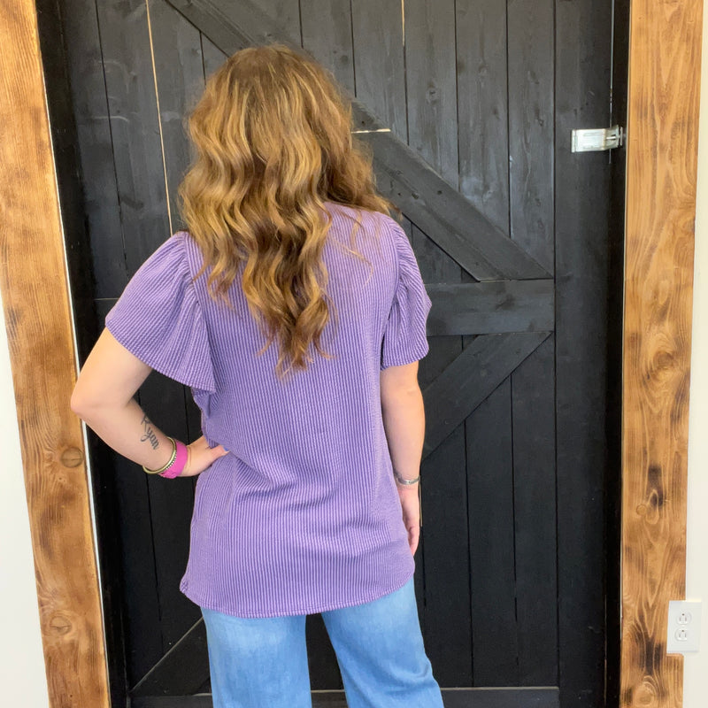 Person wearing a purple top and blue jeans standing in front of a wooden door.