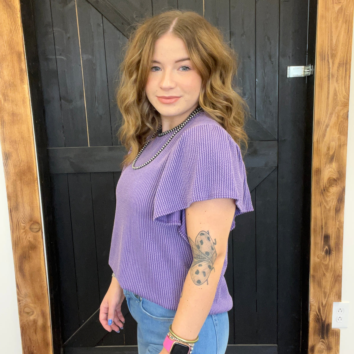 Woman wearing a purple top and blue jeans standing in front of a wooden door.