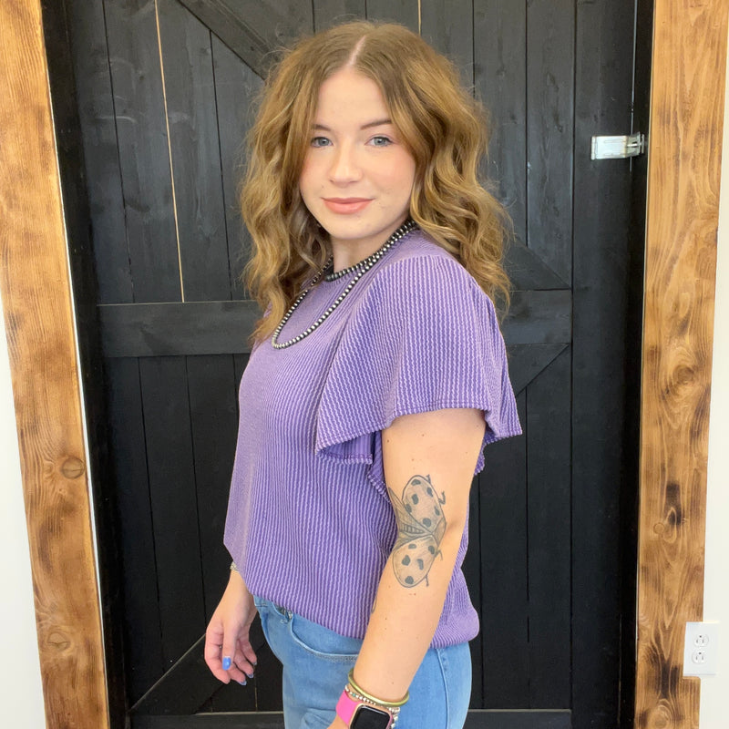 Woman wearing a purple top and blue jeans standing in front of a wooden door.