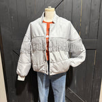 Mannequin wearing a light gray puffer jacket with fringe details against a dark wooden background.