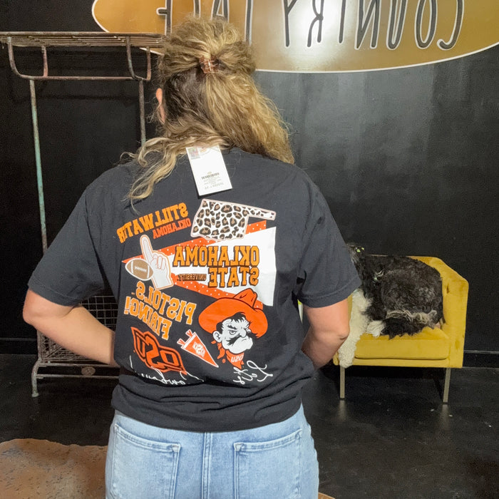 Person wearing a black t-shirt with colorful graphics and text, standing in front of a black wall with a yellow chair and decorative items.