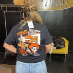 Person wearing a black t-shirt with colorful graphics and text, standing in front of a black wall with a yellow chair and decorative items.