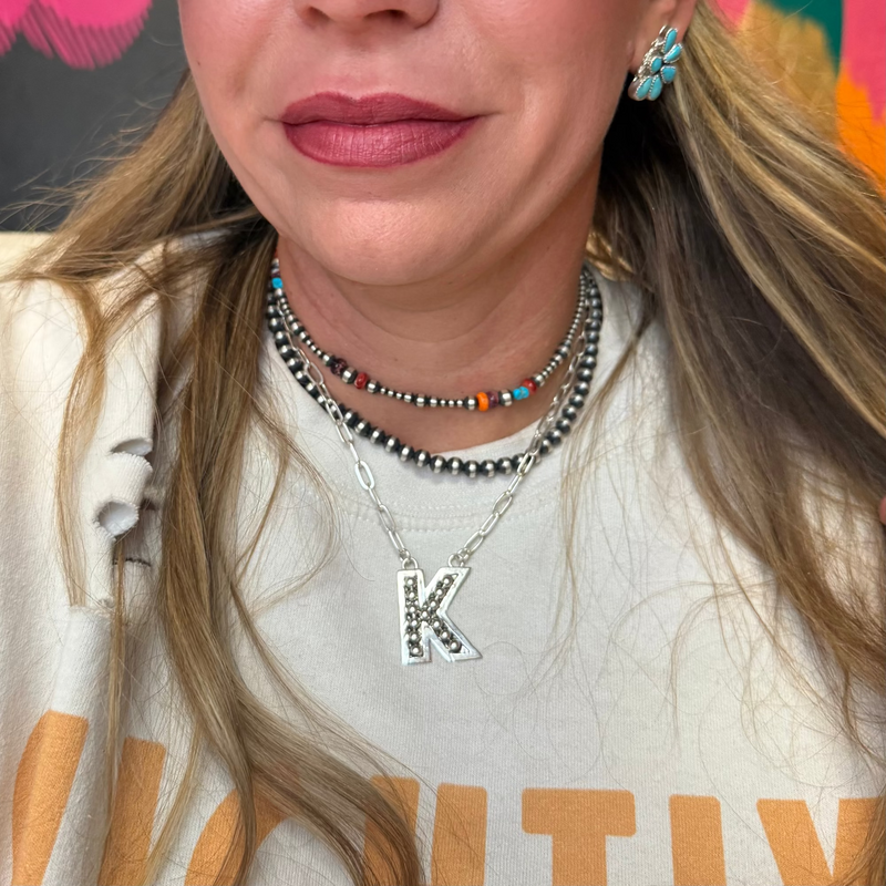 Close-up of a person wearing a necklace with a 'K' pendant, earrings, and a colorful background.