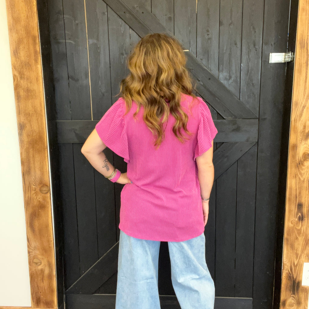 Person wearing a pink shirt and light blue jeans standing in front of a wooden door.