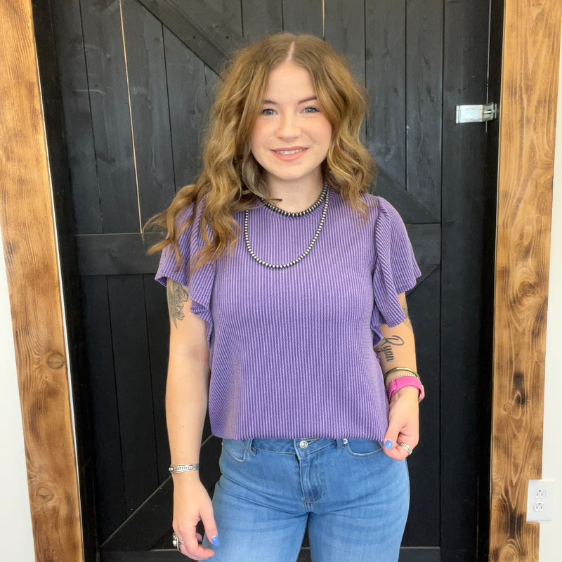 Person wearing a purple ribbed top and blue jeans standing in front of a wooden door.