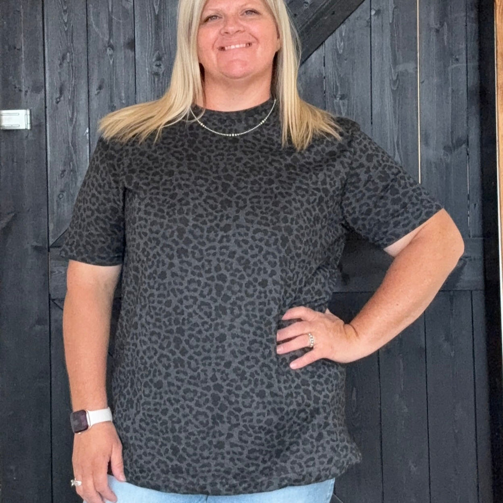 Woman wearing a black patterned top and light blue jeans standing in front of a dark wooden door.
