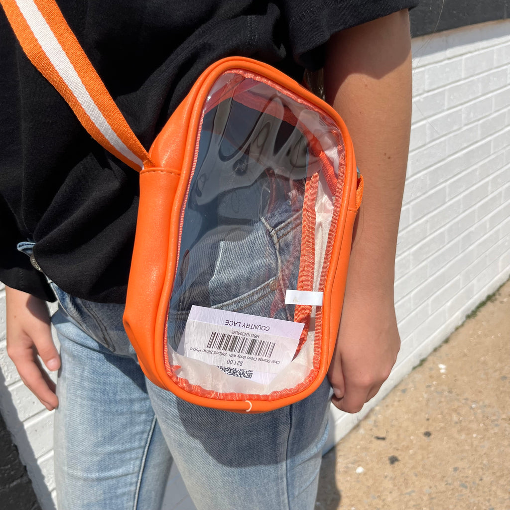 Person wearing an orange crossbody bag with a clear front pocket, standing against a white brick wall.