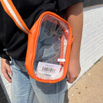 Person wearing an orange crossbody bag with a clear front pocket, standing against a white brick wall.