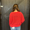 Person wearing a red sweater and blue jeans standing in front of a wooden door.