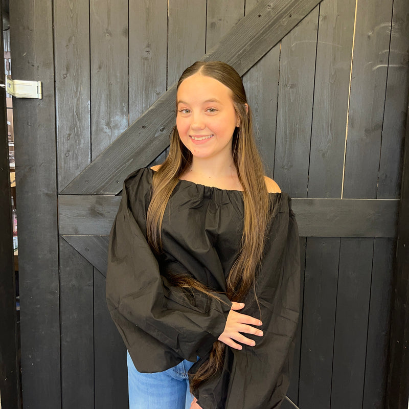 Young woman wearing a black off-shoulder top and blue jeans standing against a wooden door.