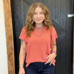 Woman wearing a coral top and dark jeans standing in front of a wooden door.