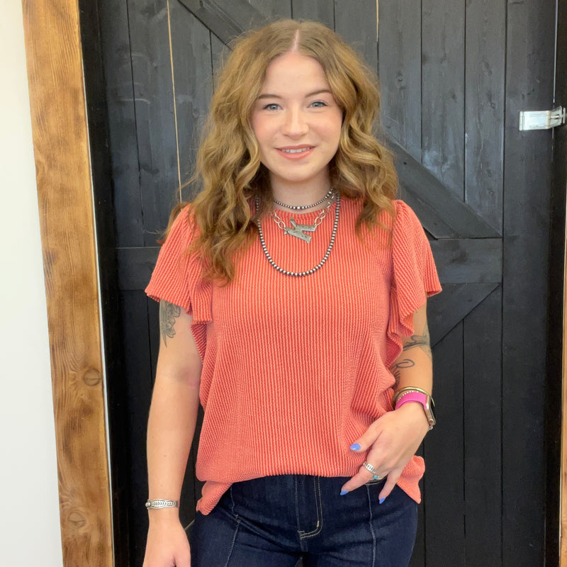 Woman wearing a coral top and dark jeans standing in front of a wooden door.