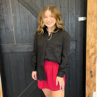 Woman wearing a black shirt and red shorts standing in front of a wooden door.