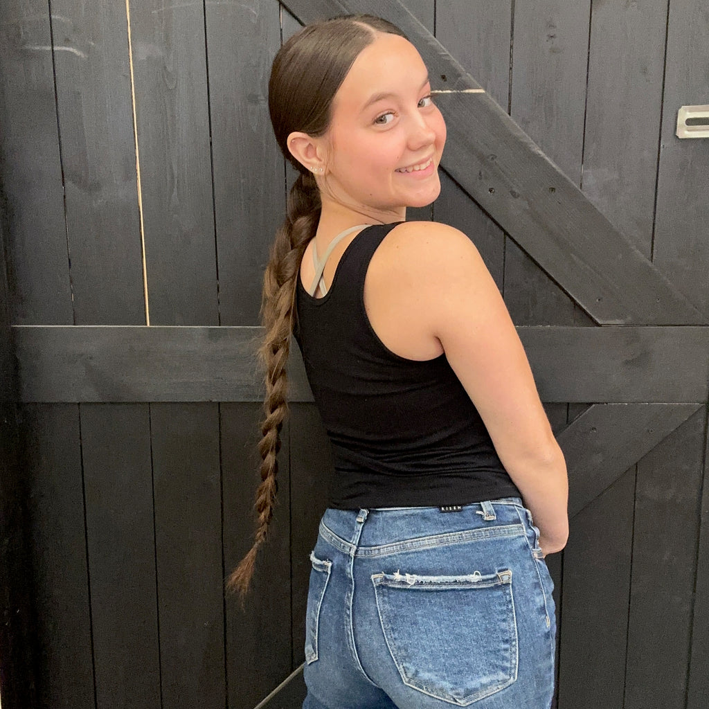 black tank top and blue jeans standing against a dark wooden door.