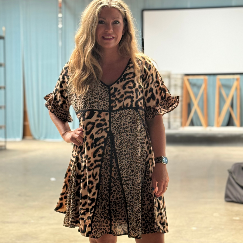 Woman wearing a leopard print dress in an indoor setting with a blue curtain and wooden beams.
