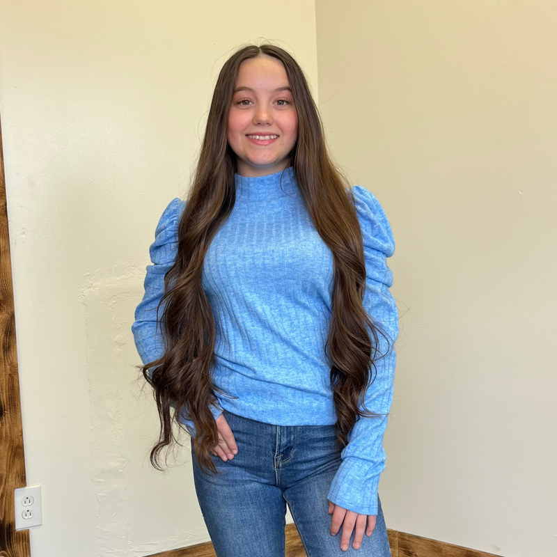 Woman wearing a blue sweater and jeans standing against a beige wall.