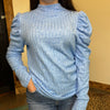 Person wearing a blue textured long-sleeve top with ruffled shoulders against a neutral background.