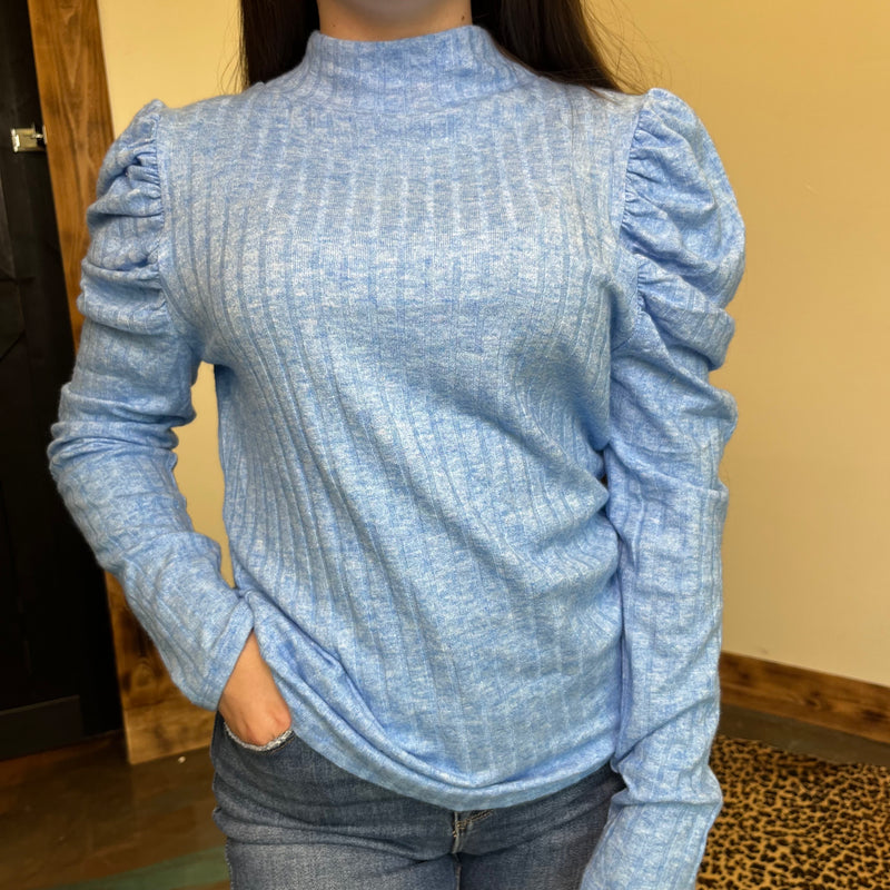 Person wearing a blue textured long-sleeve top with ruffled shoulders against a neutral background.