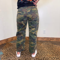 camouflage pants and a black shirt with orange text, standing on a leopard print rug.