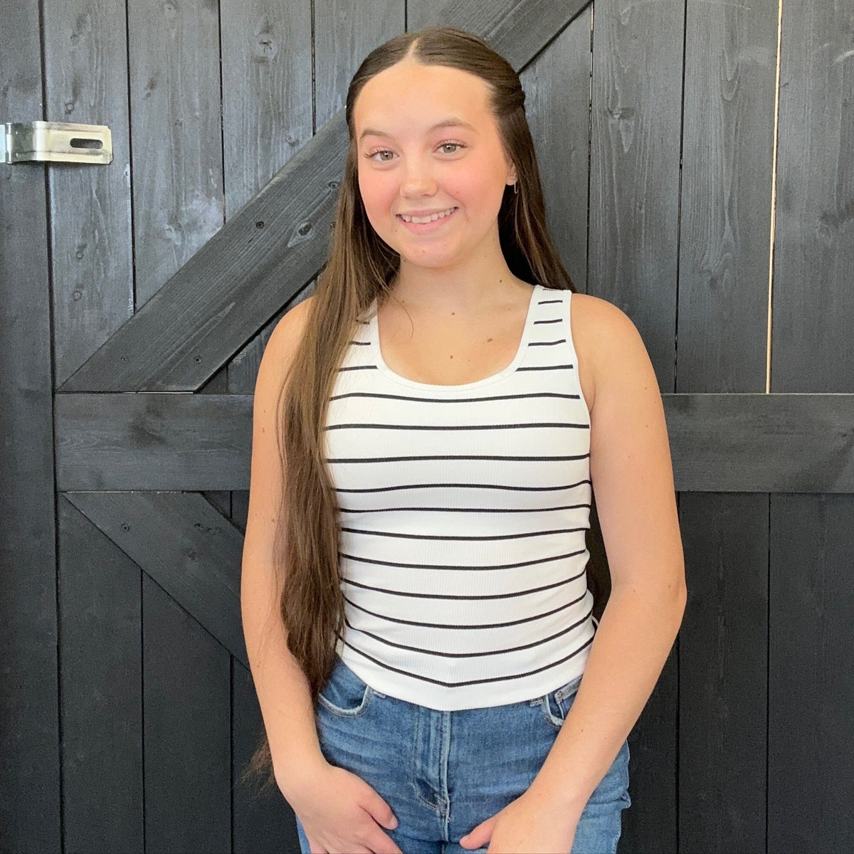 Young girl wearing a striped tank top and jeans standing in front of a wooden door.