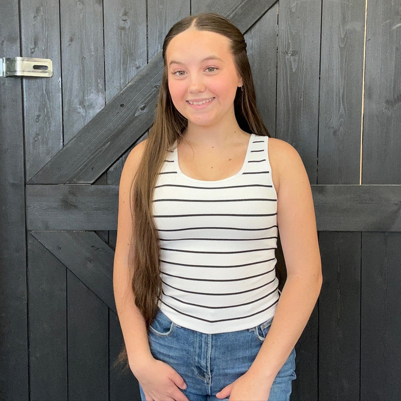 Young girl wearing a striped tank top and jeans standing in front of a wooden door.